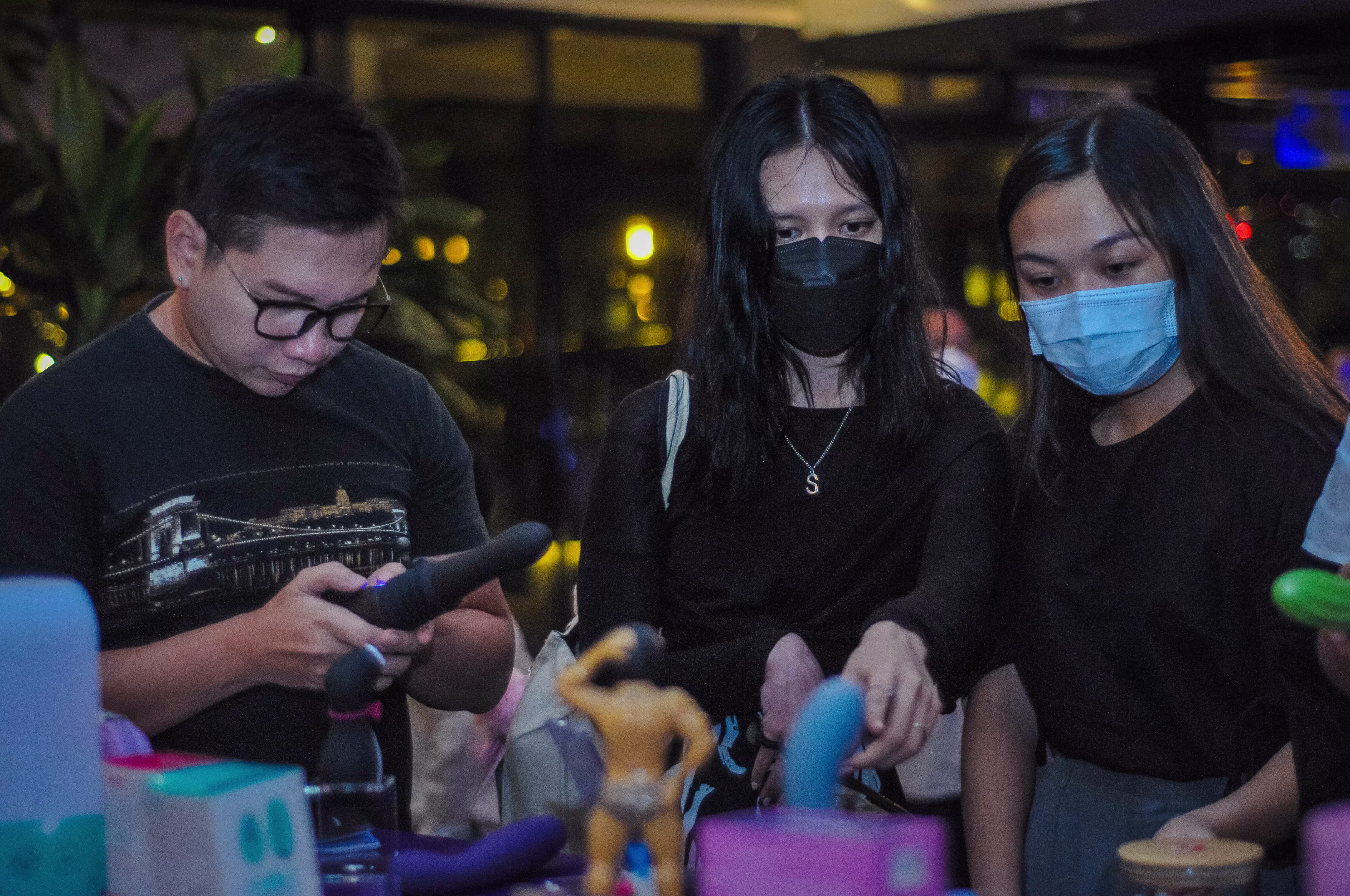 Three guests exploring the exhibition, with two masked women viewing the product display table and a man examining a vibrator