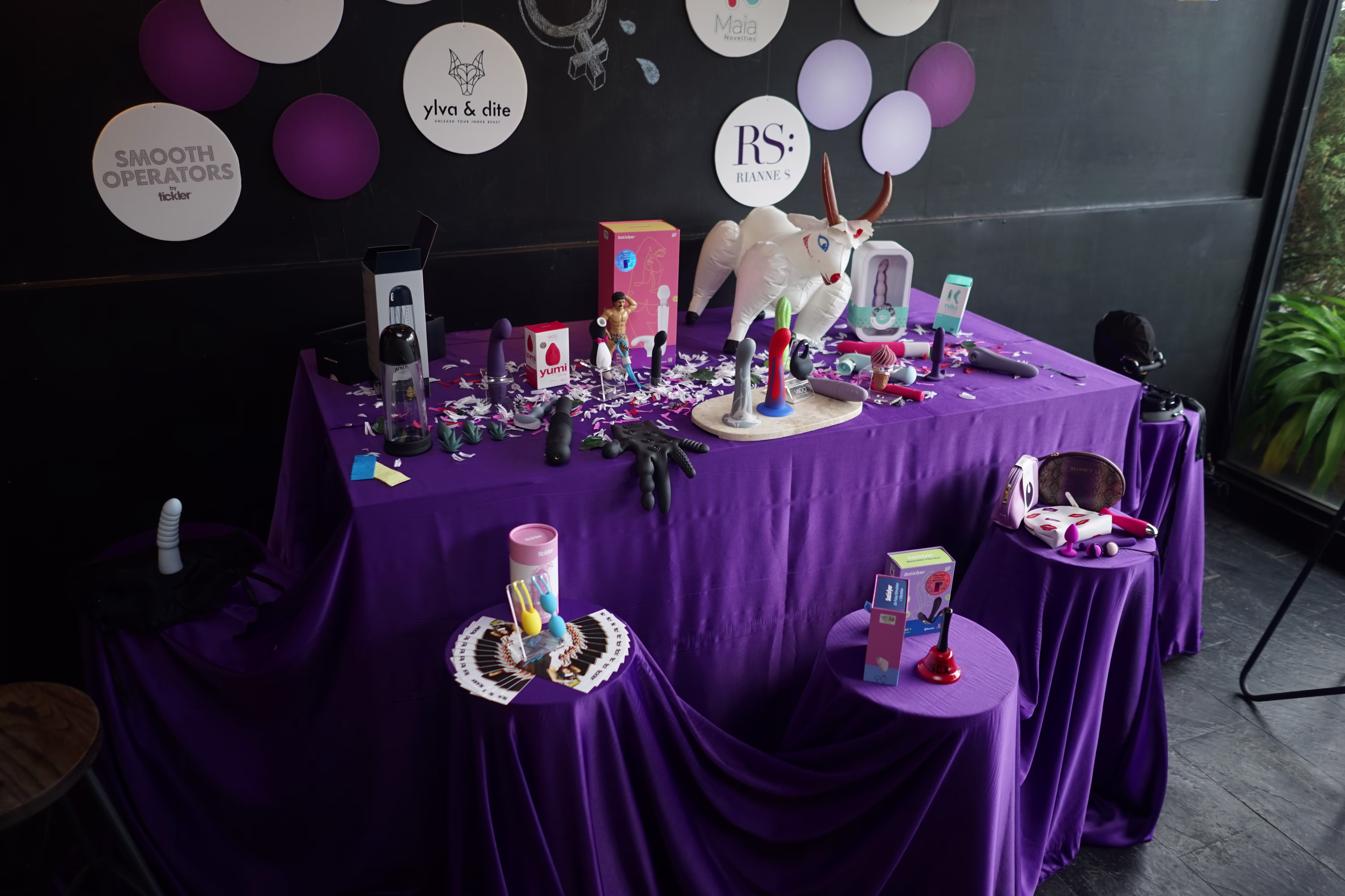 A side-profile shot of the sexual wellness product display table with a purple cloth, showcasing items against a wall featuring branded logo boards at the Laciasmara x Angsa Merah Klinik joint press conference